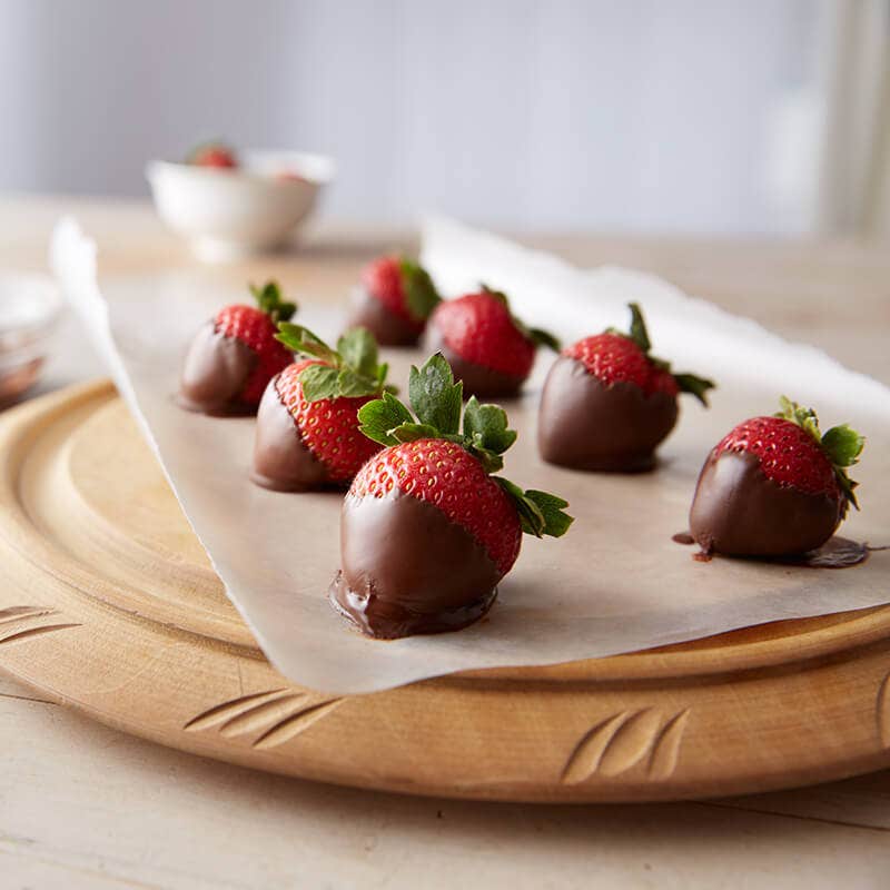 seven chocolate covered strawberries sitting on wax paper on a light brown cutting board with bowls in the background. 