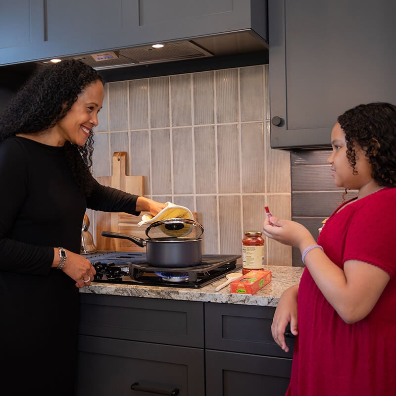 Adult and child cooking pasta in a kithen with dark cabinets and tan colored countertop.