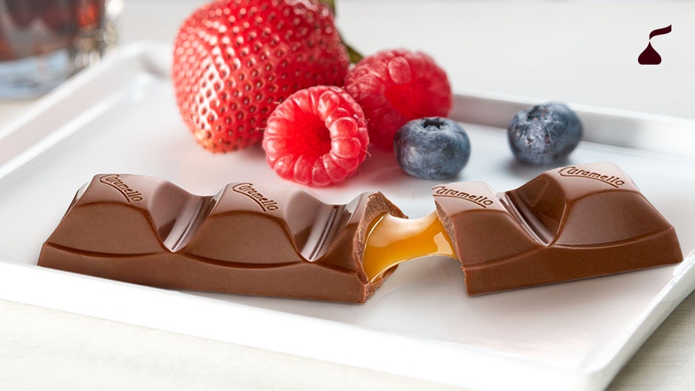A Cadbury Caramello bar broken in half with caramel stretching between the pieces on a white plate with fresh berries